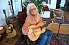 Arlo Guthrie sits in a red chair playing an acoustic guitar in his home.