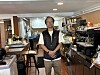 A person wearing glasses and a dark vest stands in a well-equipped café kitchen.