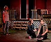 Three performers pose on a rustic stage beside railings, stacked dishes, and a wash basin.